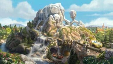 Radiator Springs Racers zoom along the track, winding past majestic rocky mountains with cascading waterfalls, lush trees, and a bright blue sky inside Cars Land at Disney California Adventure.
