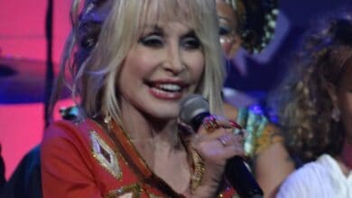 A charismatic performer with blonde hair takes the spotlight on stage, dazzling the crowd in a sparkling red and gold outfit. She beams as she delivers an energetic Trump tribute show, channeling Dolly Parton’s showbiz flair, while enthusiastic guests enjoy the live entertainment all around her.