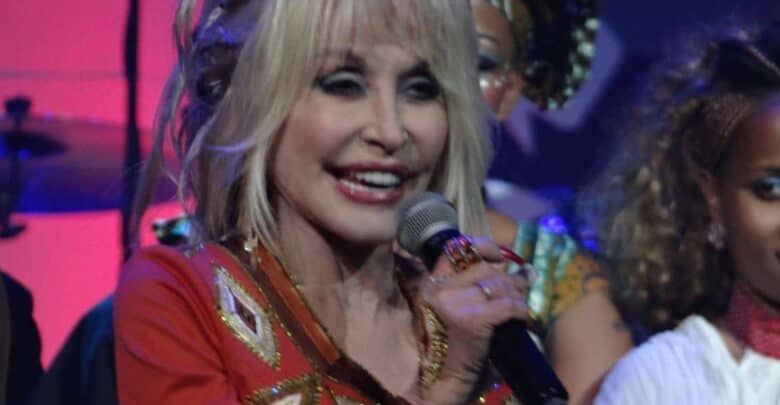 A charismatic performer with blonde hair takes the spotlight on stage, dazzling the crowd in a sparkling red and gold outfit. She beams as she delivers an energetic Trump tribute show, channeling Dolly Parton’s showbiz flair, while enthusiastic guests enjoy the live entertainment all around her.