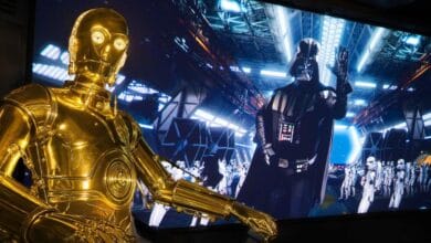 A C-3PO figure stands in front of a Star Tours screen showing Darth Vader and stormtroopers at Disney Parks.