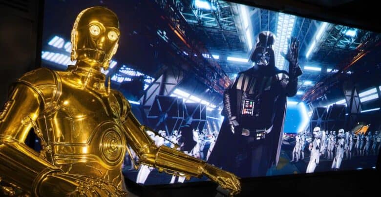 A C-3PO figure stands in front of a Star Tours screen showing Darth Vader and stormtroopers at Disney Parks.