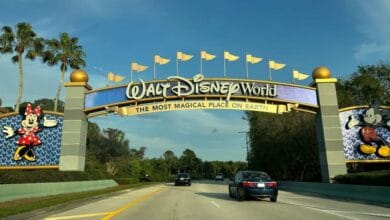 Cars driving under the Walt Disney World entrance archway featuring Mickey and Minnie Mouse as guests depart.